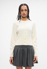 Cardigã de malha em marfim com decote redondo, fecho de botão e textura canelada. Combinado com uma saia cinza com riscas à medida.