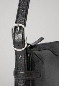 Cinghia nera in pelle con superficie testurizzata, fibbia in metallo argentato e fori per la regolazione. La cinghia è attaccata al lato della borsa con hardware metallico.