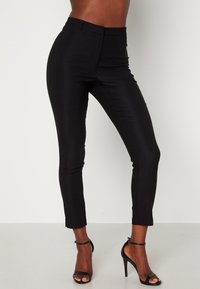 Pantalon noir taille haute avec une coupe ajustée et une texture lisse, doté d'une jambe fuselée et de fentes discrètes aux chevilles.