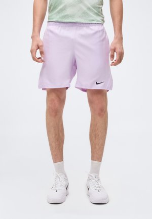 Mann iført lyse lilla Nike treningsshorts, hvite Nike joggesko og hvite sokker, stående mot en enkel lys bakgrunn.