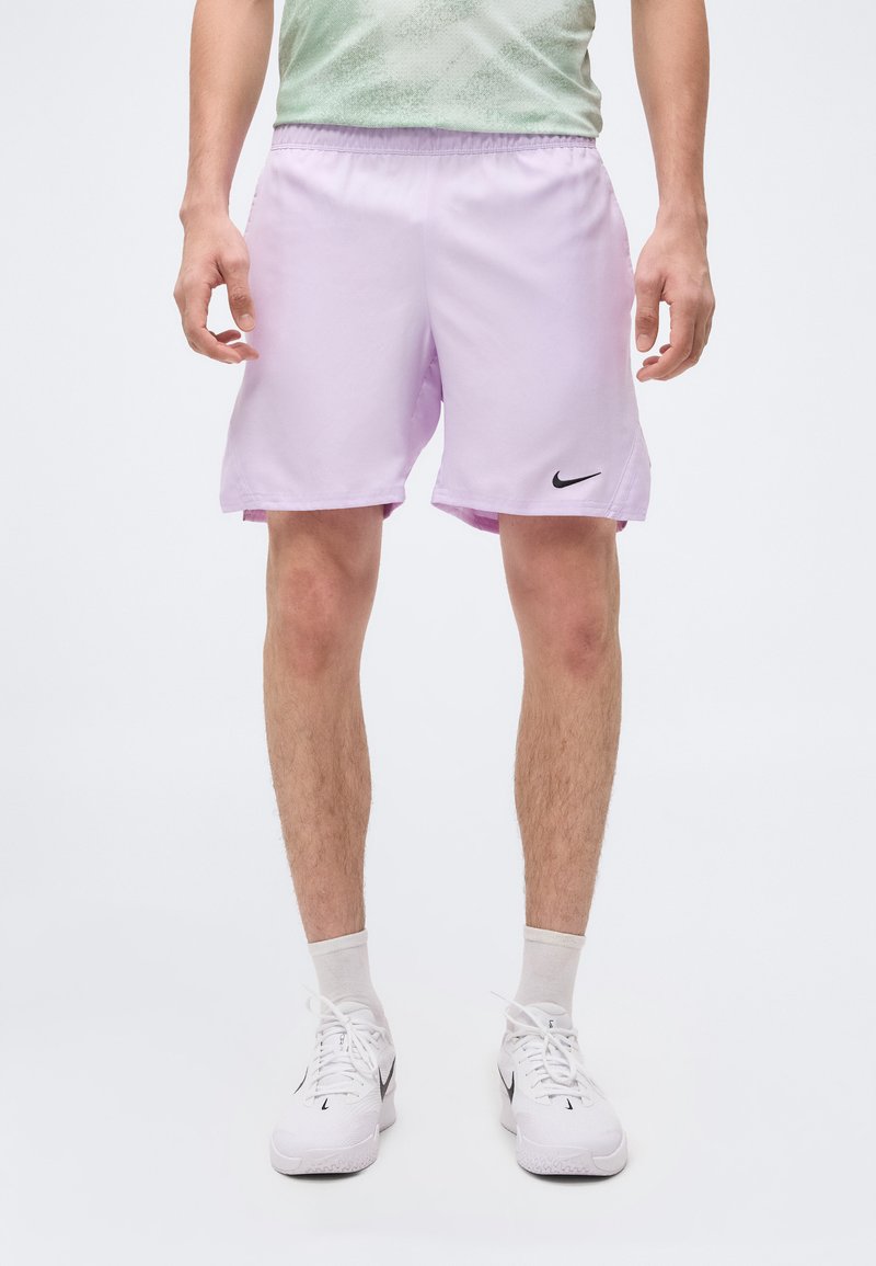 Homme portant un short de sport Nike violet clair, des baskets Nike blanches et des chaussettes blanches, debout devant un fond clair uni.