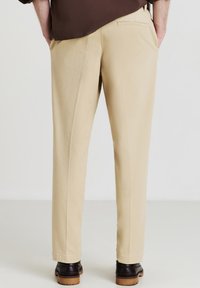 Uomo che indossa pantaloni beige a vestibilità dritta e scarpe di pelle nere, in piedi con le mani nelle tasche, visto da dietro.