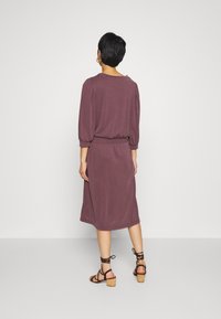 Mörkviolett klänning med trekvartslång ärm, rynkad axeldetalj, elastisk midja och nedre del som når till vaden, i kombination med bruna sandaler med remmar.