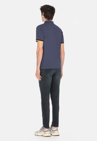 Polo shirt navy con accenti gialli e neri sui polsini, abbinato a jeans scuri e sneakers grigie, visto da dietro.