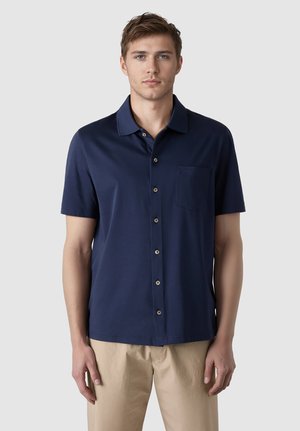 Młody mężczyzna stojący przodem, ubrany w granatową koszulę na krótki rękaw zapinaną na guziki oraz beżowe spodnie, na tle jednolitego szarego tła.