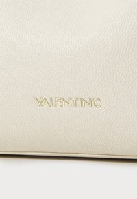Kreminės spalvos odinė rankinė su akmeniniu tekstūru, su iškilnotu auksiniu „VALENTINO“ logotipu priekyje. Paprastas, stačiakampio formos dizainas.