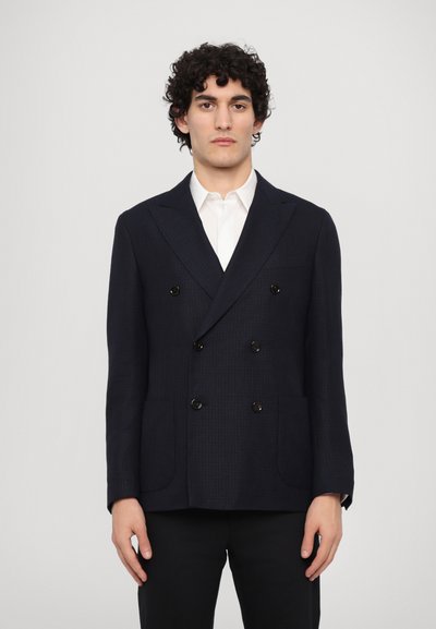 Blazer marine à double boutonnage en tissu texturé, avec deux poches avant et des boutons noirs, associé à une chemise blanche en dessous.