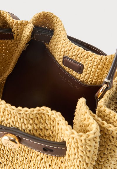 Ανοιχτή υφαντή τσάντα από άχυρο με καφέ δερμάτινη επένδυση, κούμπωμα με χρυσό κουμπί και εμφανές όνομα μάρκας στο εσωτερικό.