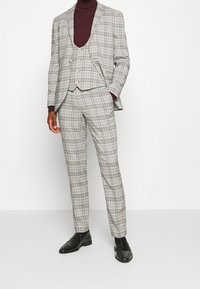 Grijs geruit pak met een getailleerde jas, bijpassende broek en een bordeauxrode coltrui eronder. Zwarte dresschoenen maken de look compleet.