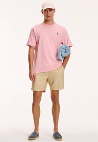 Rózsaszín rövid ujjú póló kis kék logóval, bézs rövidnadrág és kék törölköző; viselve világos rózsaszín sapkával és kék espadrille cipővel.