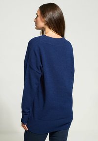 Frau mit langen braunen Haaren, die goldene Ohrringe und einen locker sitzenden dunkelblauen Pullover trägt, von hinten vor einem schlichten Hintergrund gesehen.