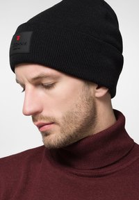 Czarna dziana czapka beanie z czarną prostokątną naszywką z czerwonym tekstem, noszona na bordowym swetrze z golfem. Posiada prążkowaną teksturę.