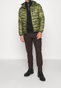 Olivengrøn pufferjakke med hætte, lynlås-lukning og kviltet design, parret med mørkebrune bukser og sorte snørebåndsstøvler.