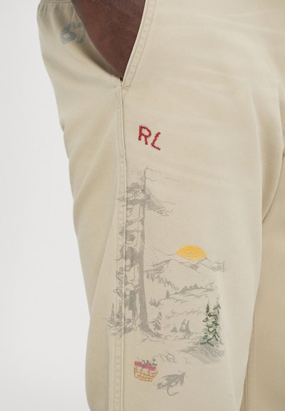 Calças bege com paisagem bordada que inclui um sol, montanhas, árvores e uma cesta de frutas. "RL" bordado em vermelho.