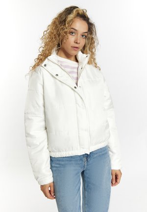 Ung kvinde med krøllet hår iført en hvid dunjakke, stribet skjorte og blå jeans mod en ensfarvet hvid baggrund.