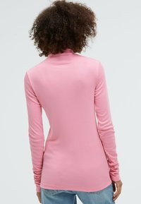Long-sleeve, fitted turtleneck in light pink, made of soft, stretchy fabric. Features a high collar and smooth texture, paired with denim jeans.