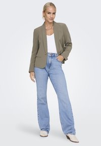Veste vert olive avec un col cranté, associée à un haut blanc et un jean évasé bleu clair. Le modèle porte des bottes à talons beiges.