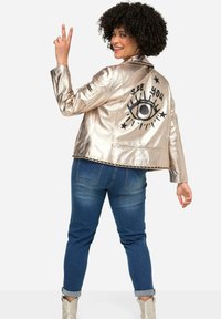 Metallic silberne Jacke mit einem schwarzen grafischen Auge und dem Text "SEE YOU" auf dem Rücken. Kombiniert mit blauen Jeans und hellen Knöchelstiefeln.