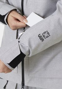 Šedá texturovaná bunda s kapsou na zip na rukávu, s černým logem. Ruka držící bílou kartu umístěná blízko kapsy.