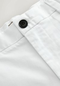 Ceinture de pantalon blanche avec un bouton noir et des détails de couture de boutonnière visibles sur un fond uni.