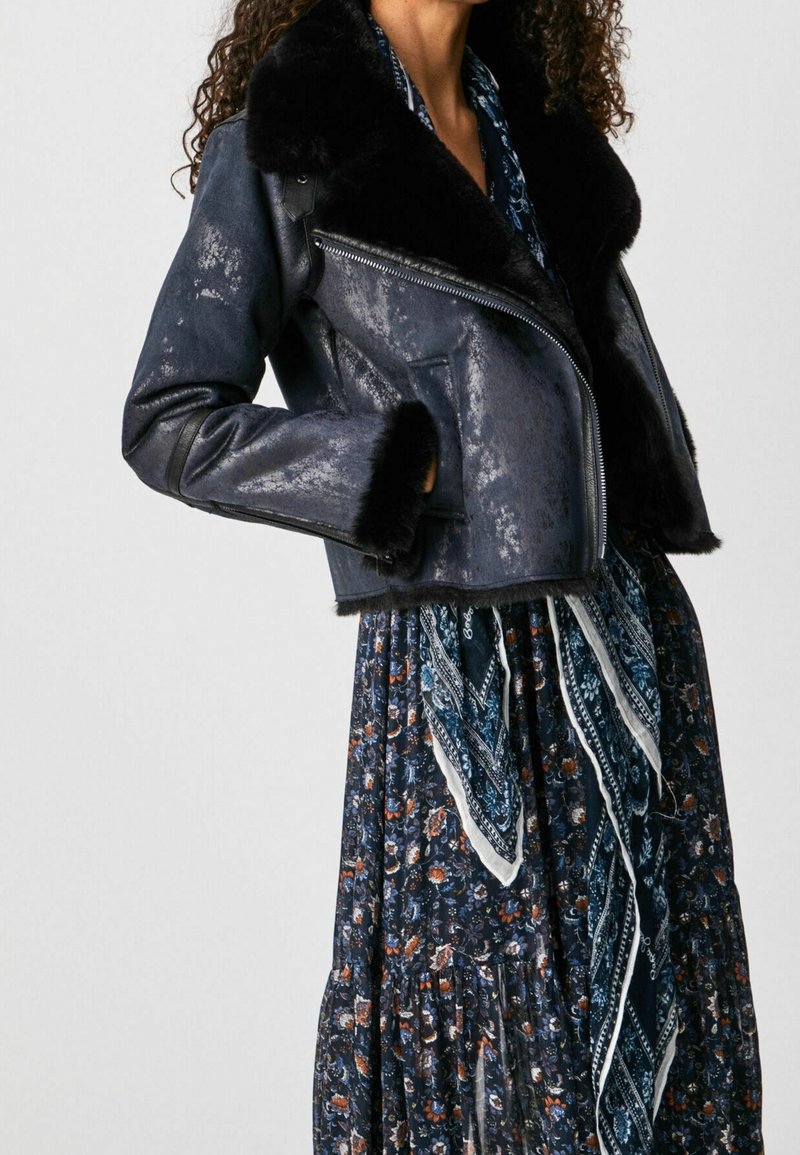 Giacca di pelle blu navy con colletto in pelliccia nera, design cropped e chiusura con zip. Abbinata a un lungo abito con motivo floreale e a una sciarpa.