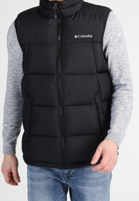 Svart vadderad väst med hög krage och framdragkedja, med en logotyp på övre vänstra sidan. Bärs över en grå långärmad tröja.