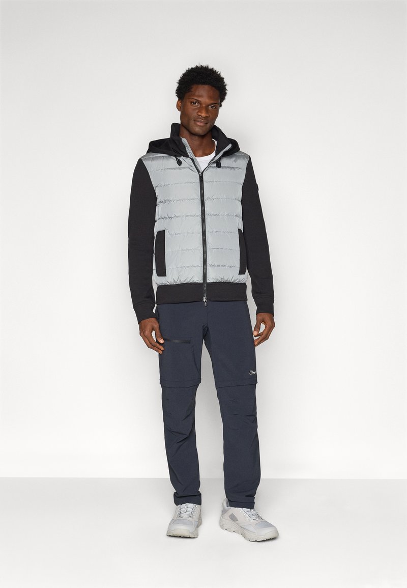 Izolowana szara kamizelka puchowa z czarnymi rękawami i kapturem, z przodu na zamek błyskawiczny, w połączeniu z ciemnymi spodniami cargo i jasnoszarymi trampkami.