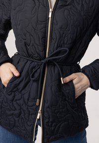 Veste matelassée navy avec un design structuré, une fermeture éclair dorée, une taille à cordon de serrage et des poches latérales, présentant un motif texturé en relief.