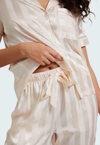 Satin pajama set in cream with soft stripes. Short-sleeve shirt features a pocket and buttons; pants with elastic waistband and ribbon tie.