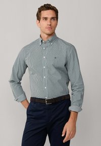 Camisa de botones a cuadros verdes y blancos, de manga larga, con cuello y un pequeño logotipo. Combinada con pantalones azul marino oscuro y un cinturón oscuro.