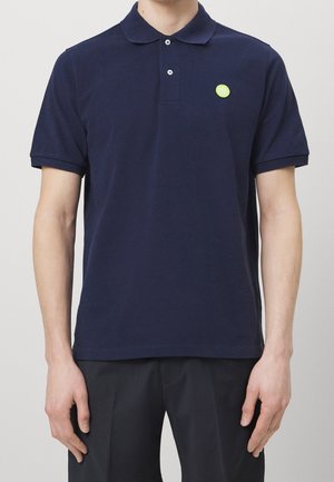 Mann trägt ein marineblaues kurzärmeliges Poloshirt mit zwei weißen Knöpfen und einem kleinen runden gelben Logo auf der linken Brust.