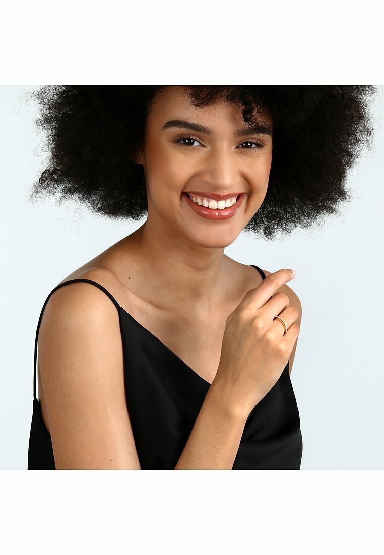 Guld ring på en hand med något böjda fingrar, bärs av en person i en svart topp. Bakgrunden är ljus och neutral, vilket framhäver ringen.