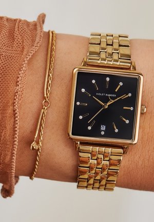 Montre carrée dorée avec un cadran noir, marqueurs d'heure en diamants, aiguilles dorées et affichage de la date. Portée sur un poignet avec un bracelet en chaîne doré.