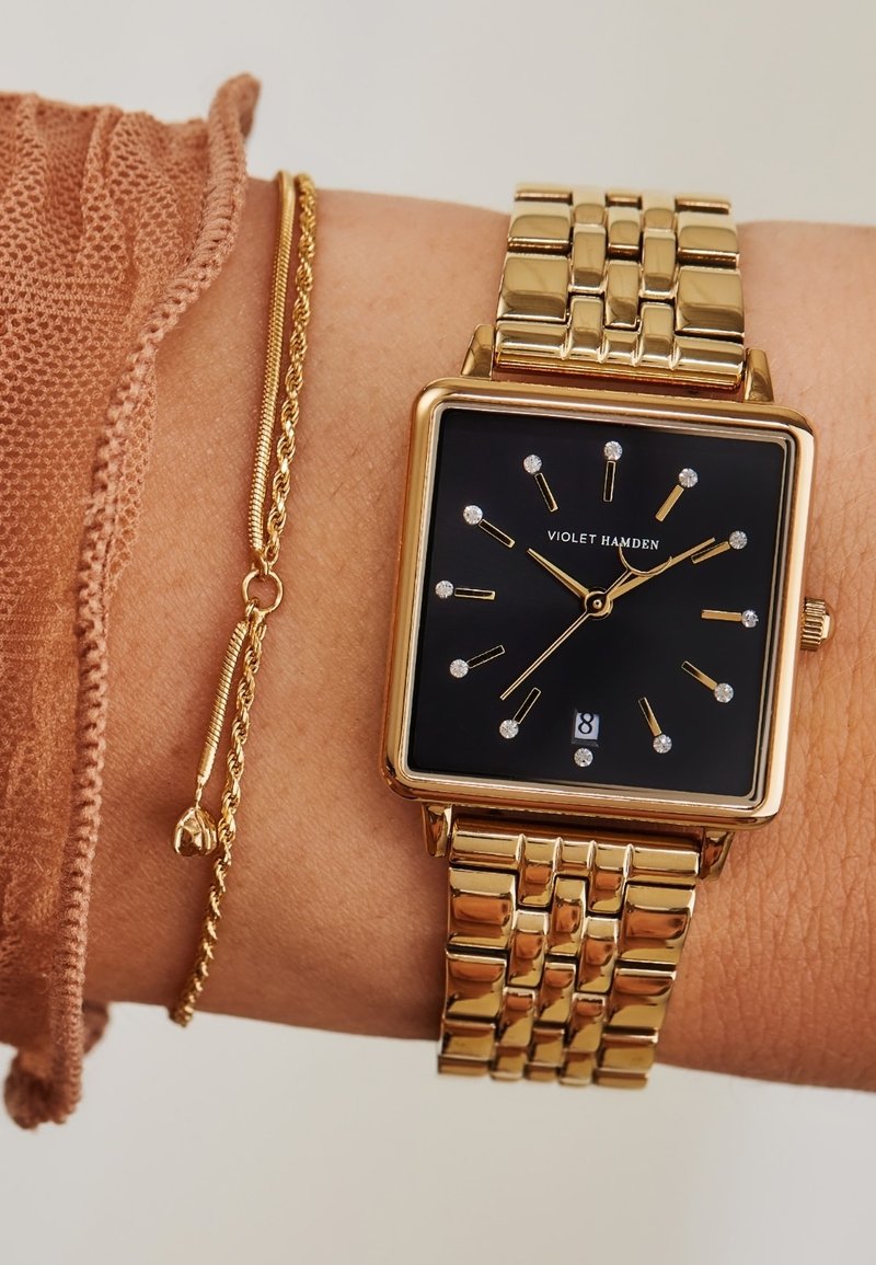 Goldener quadratischer Uhr mit schwarzem Zifferblatt, Diamant-Stundenmarkierungen, goldenen Zeigern und Datumsanzeige. Trägt am Handgelenk mit einem goldenen Kettenarmband.