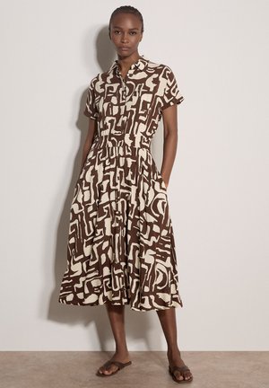 Frau trägt ein braun-cremefarbenes Hemdkleid mit abstraktem Muster, kurzen Ärmeln, Knopfleiste vorne, Taschen und braunen Pantoletten, steht an einer Wand.