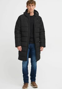 Černý péřový kabát s kapucí, prošívaná textura, plný zip, střední délka, nošený s černým tričkem a modrými džíny. Viditelné hnědé boty.