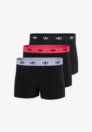 Junger Mann steht barfuß, trägt schwarze Boxer-Shorts mit einem weißen Bund, der schwarze Trefoil-Logos zeigt, vor einem einfarbigen weißen Hintergrund.