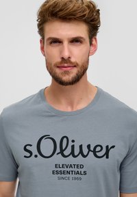 Siva pamučna majica s okruglim izrezom, s velikim crnim tiskanim logom "s.Oliver" i natpisom "ELEVATED ESSENTIALS SINCE 1969."
