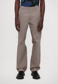 Mężczyzna w taupe prostych spodniach, czarnych sneakersach i czarnej koszuli w niebiesko-zielony kwiatowy wzór, stojący na tle białym.