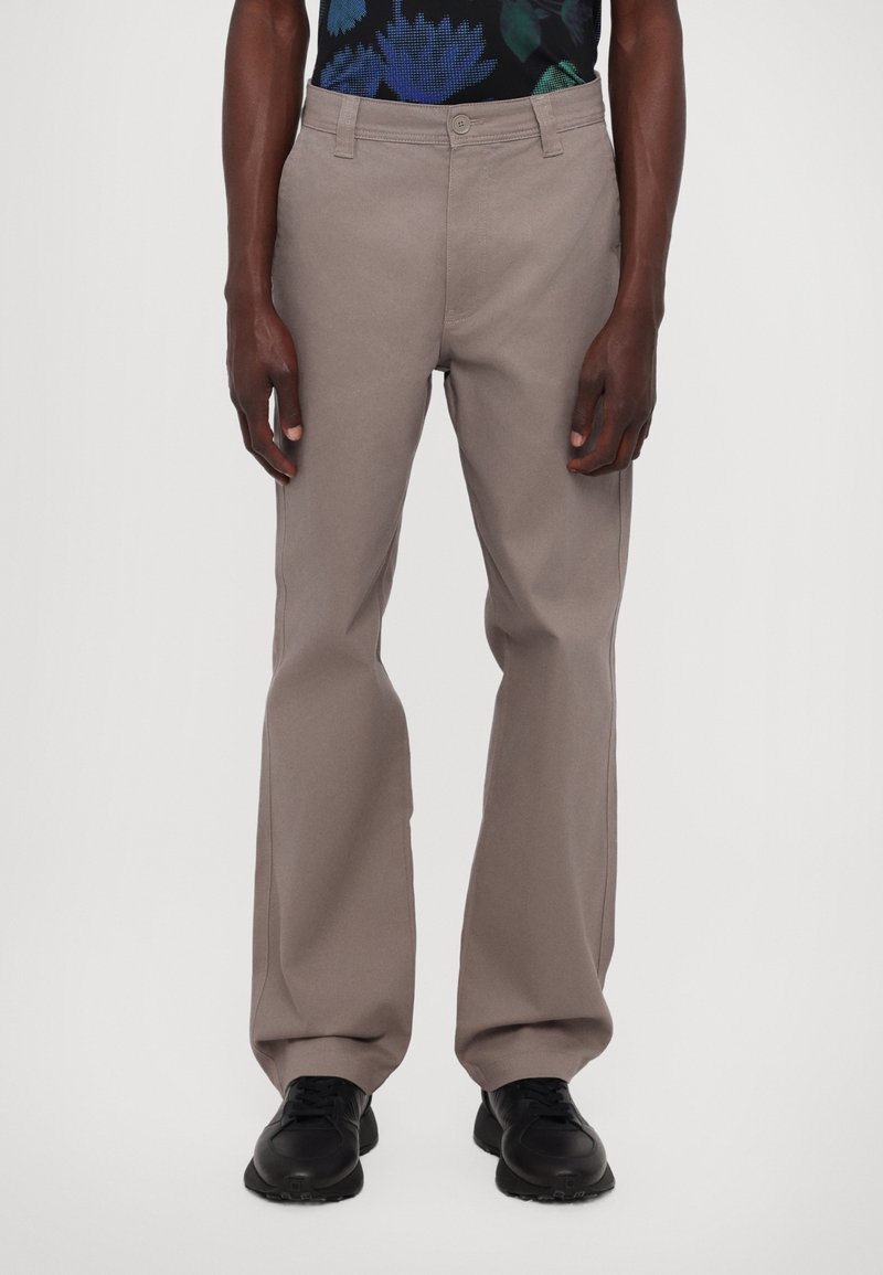 Mężczyzna w taupe prostych spodniach, czarnych sneakersach i czarnej koszuli w niebiesko-zielony kwiatowy wzór, stojący na tle białym.