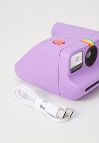 Fialový instantní fotoaparát s červeným spoušťovým tlačítkem a duhovým pruhem, vedle zkrouceného bílého USB-C nabíjecího kabelu na bílé ploše.