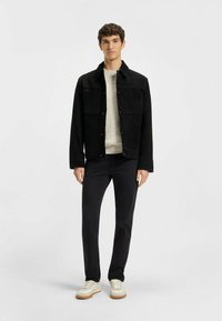 Svart mockajacka med två bröstfickor, tillsammans med en texturerad krämfärgad tröja, svarta byxor och ljusa sneakers med en tan-färgad sula.