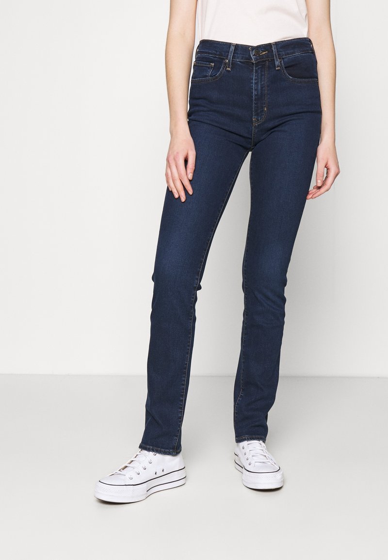 Femme portant un jean skinny bleu foncé, des baskets blanches à semelles compensées et un haut blanc rentré, debout devant un fond uni.