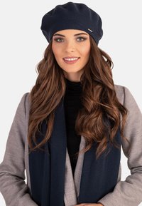 Tengerészkék kötött baret, texturált felülettel, szürke kardigánnal és sötét sállal párosítva. Hosszú, hullámos barna haj látható.