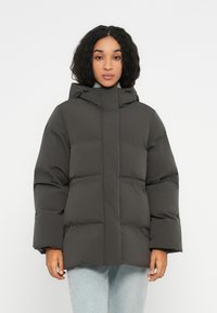 Mujer joven vestida con una chaqueta de invierno acolchada gris oscuro con capucha y vaqueros de color azul claro, de pie contra un fondo blanco liso.