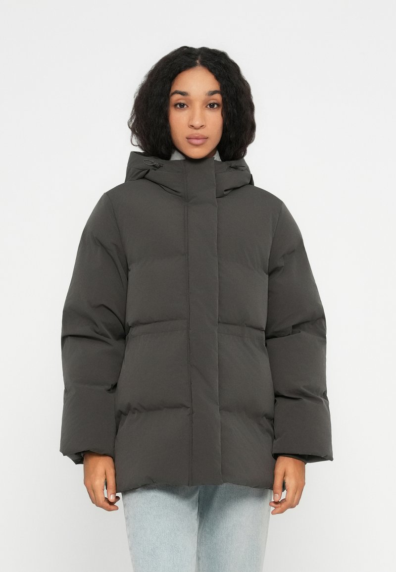 Mujer joven vestida con una chaqueta de invierno acolchada gris oscuro con capucha y vaqueros de color azul claro, de pie contra un fondo blanco liso.