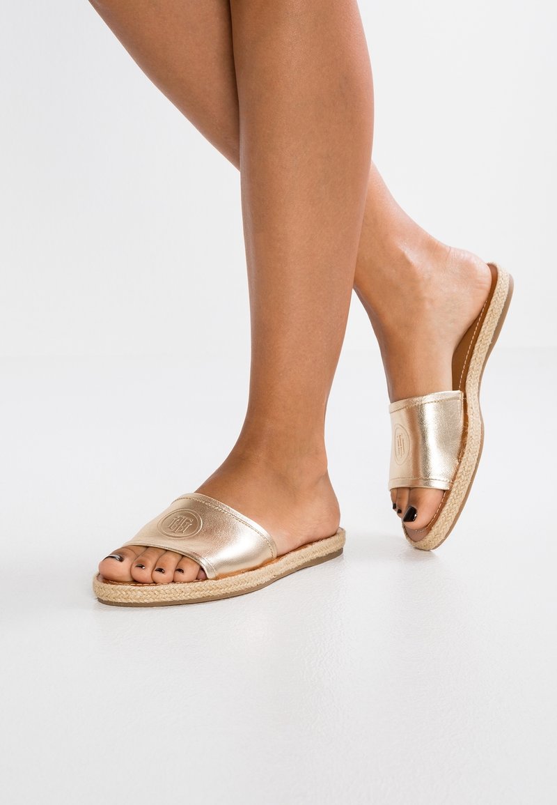 Jambes portant des sandales slides métalliques dorées avec des semelles tissées, orteils peints en noir, debout sur une surface blanche et lisse.