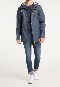 Veste imperméable bleue avec capuche, dotée d'accents jaunes, de poches latérales et d'une fermeture éclair. Portée par-dessus une chemise à motif sombre et un jean.