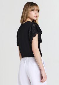 Giovane donna che si volta, indossando un top nero con maniche corte in pizzo e pantaloni bianchi larghi, davanti a uno sfondo neutro.