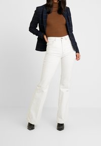Jean flare blancs avec un design à cinq poches, taille haute et détails de couture minimaux, associés à un col roulé côtelé brun et un blazer à carreaux bleu marine.